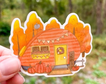 May include: A sticker of a red and white striped camper with a yellow door and a wreath. The camper has a window with flowers and a sign that says "Welcome". There are pumpkins in front of the camper and trees with orange leaves behind it.