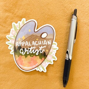 May include: A white sticker with a blue, green, and brown landscape scene on a paint palette. The text "Appalachian artist" is written in black cursive. The sticker has a white border and pink flowers around the edges. The website "www.jammamafoundation.com" is printed at the bottom of the sticker.