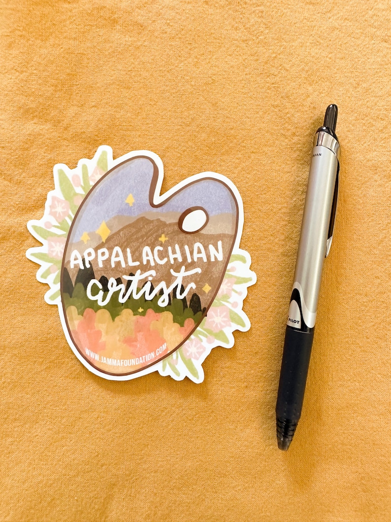 May include: A white sticker with a blue, green, and brown landscape scene on a paint palette. The text "Appalachian artist" is written in black cursive. The sticker has a white border and pink flowers around the edges. The website "www.jammamafoundation.com" is printed at the bottom of the sticker.