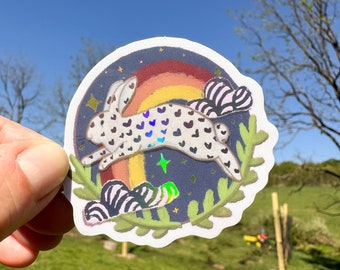 May include: A white rabbit with black spots and hearts printed on its fur leaps over a rainbow with a starry night sky background. The sticker is surrounded by green leaves and a white border.