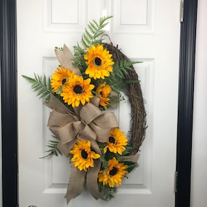 May include: A decorative oval-shaped wreath featuring bright yellow sunflowers with brown centers, green fern leaves, and a large burlap bow. The wreath is made of woven twigs and is designed for door or wall decor.