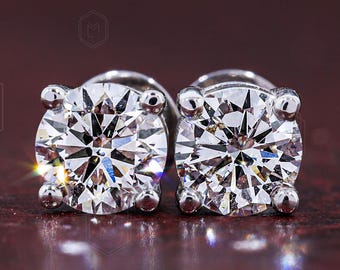 May include: A pair of round-cut stud earrings featuring clear, faceted stones set in a four-prong silver-toned metal setting. The stones exhibit high brilliance and light reflection, showcasing a classic circular design with a polished metallic finish.