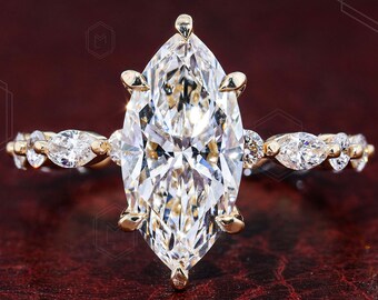 May include: A gold ring featuring a large marquise-cut center stone held by six prongs. The band is accented with smaller marquise-cut stones on each side, creating a symmetrical design. The metal has a polished finish and a warm yellow gold tone.