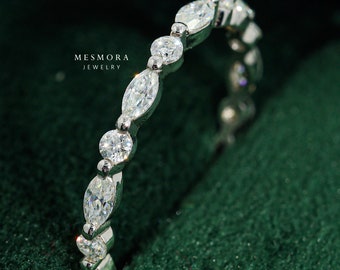 Puede incluir: Un anillo de banda en tono plateado con un patrón alterno de piedras transparentes de talla marquesa y redonda engastadas en metal. El texto MESMORA JEWELRY es visible cerca del anillo. El diseño muestra una secuencia de piedras facetadas.