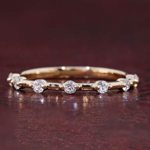 Puede incluir: Un anillo de banda dorada con una serie de piedras redondas, transparentes y facetadas, espaciadas uniformemente a lo largo de la mitad superior de la banda. El metal tiene un acabado pulido y las piedras están aseguradas en engastes.