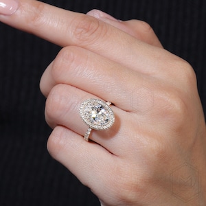 May include: A gold ring with a large oval-shaped diamond surrounded by a halo of smaller diamonds. The ring has a pave setting with smaller diamonds on the band.
