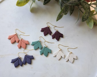 May include: A collection of handmade earrings in various colors, including peach, burgundy, teal, navy, and white. Each pair features a leaf-shaped design with gold-colored ear wires. The earrings are displayed on a white surface.