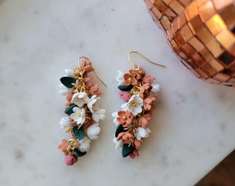Puede incluir: Un par de pendientes colgantes con racimos de pequeñas flores esculpidas en tonos terracota, blanco y rosa empolvado, acentuados con hojas de color verde oscuro y fornituras de metal dorado, suspendidos de ganchos dorados.