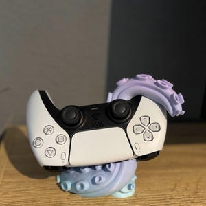 Könnte beinhalten: Ein weiß-schwarzer PlayStation 5-Controller, der auf einem hellblau-violetten, oktopusförmigen Ständer ruht. Der Controller hat schwarze Tasten und ein Steuerkreuz, mit den PlayStation-Symbolen. Der Oktopus-Ständer hat einen Farbverlauf.