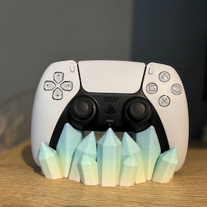 Könnte beinhalten: Ein weißer PlayStation 5-Controller ruht auf einem dekorativen Ständer. Der Ständer hat eine Reihe von stilisierten, farbverlaufenden Kristallen in Blau-, Grün- und Gelbtönen. Der Controller hat schwarze Akzente und Tasten.