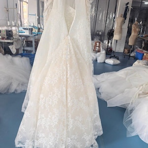 May include: A floor-length, strapless gown featuring a cream-coloured base layer overlaid with intricate white floral lace. The garment has a sweetheart neckline, a fitted bodice, and a flared skirt with a scalloped lace hemline at the bottom.