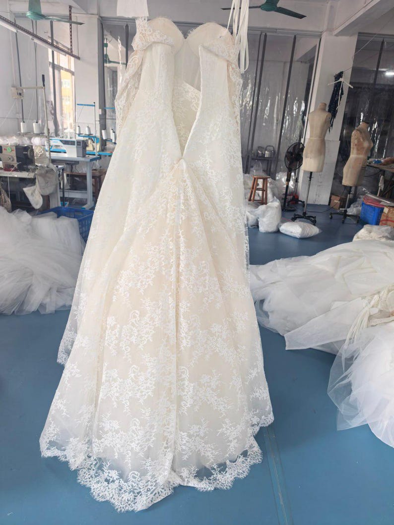 May include: A floor-length, strapless gown featuring a cream-coloured base layer overlaid with intricate white floral lace. The garment has a sweetheart neckline, a fitted bodice, and a flared skirt with a scalloped lace hemline at the bottom.