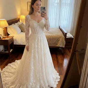 May include: A floor-length white wedding dress featuring long lace sleeves, a sweetheart neckline, and a structured corset-style bodice. The gown is adorned with intricate floral lace embroidery throughout, flowing into a full skirt with a train.