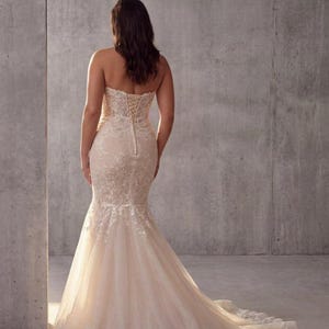 May include: A strapless, floor-length mermaid gown in a soft champagne hue, featuring intricate white floral lace overlays, a fitted bodice with a corset-style lace-up back closure, and a flared skirt that extends into a sweeping train.