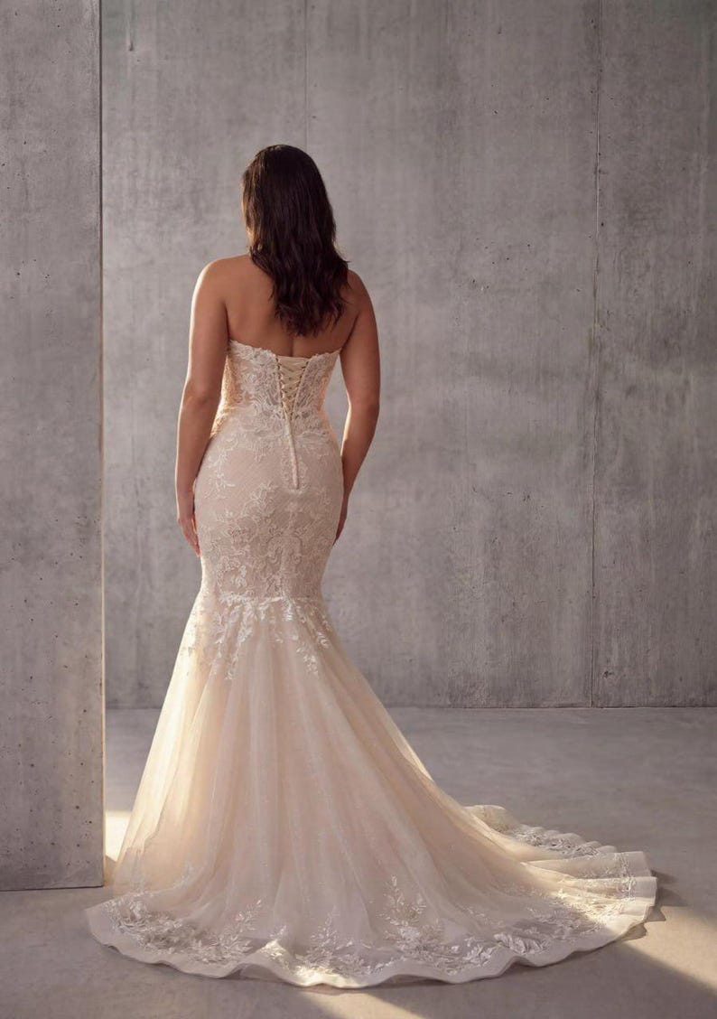 May include: A strapless, floor-length mermaid gown in a soft champagne hue, featuring intricate white floral lace overlays, a fitted bodice with a corset-style lace-up back closure, and a flared skirt that extends into a sweeping train.