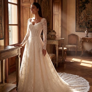 May include: A floor-length ivory lace gown featuring long sleeves, a sweetheart neckline, and a structured corset-style bodice. The dress has a full skirt with intricate floral lace detailing and a sweeping train that extends across the floor.