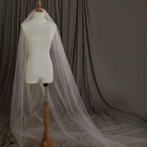 May include: A long, sheer white bridal veil made of fine tulle fabric, featuring delicate, shimmering beadwork along the entire length of the edges. The veil is draped over a mannequin, extending downwards to create a soft, flowing silhouette.