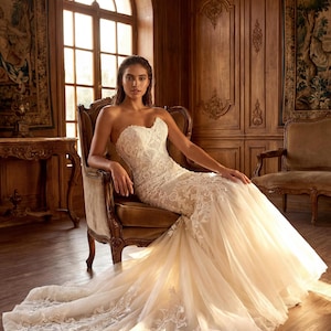 May include: A strapless ivory wedding dress featuring a sweetheart neckline and a fitted bodice covered in intricate white lace embroidery. The skirt transitions into a voluminous tulle train with matching lace appliqu&eacute;s along the hemline.