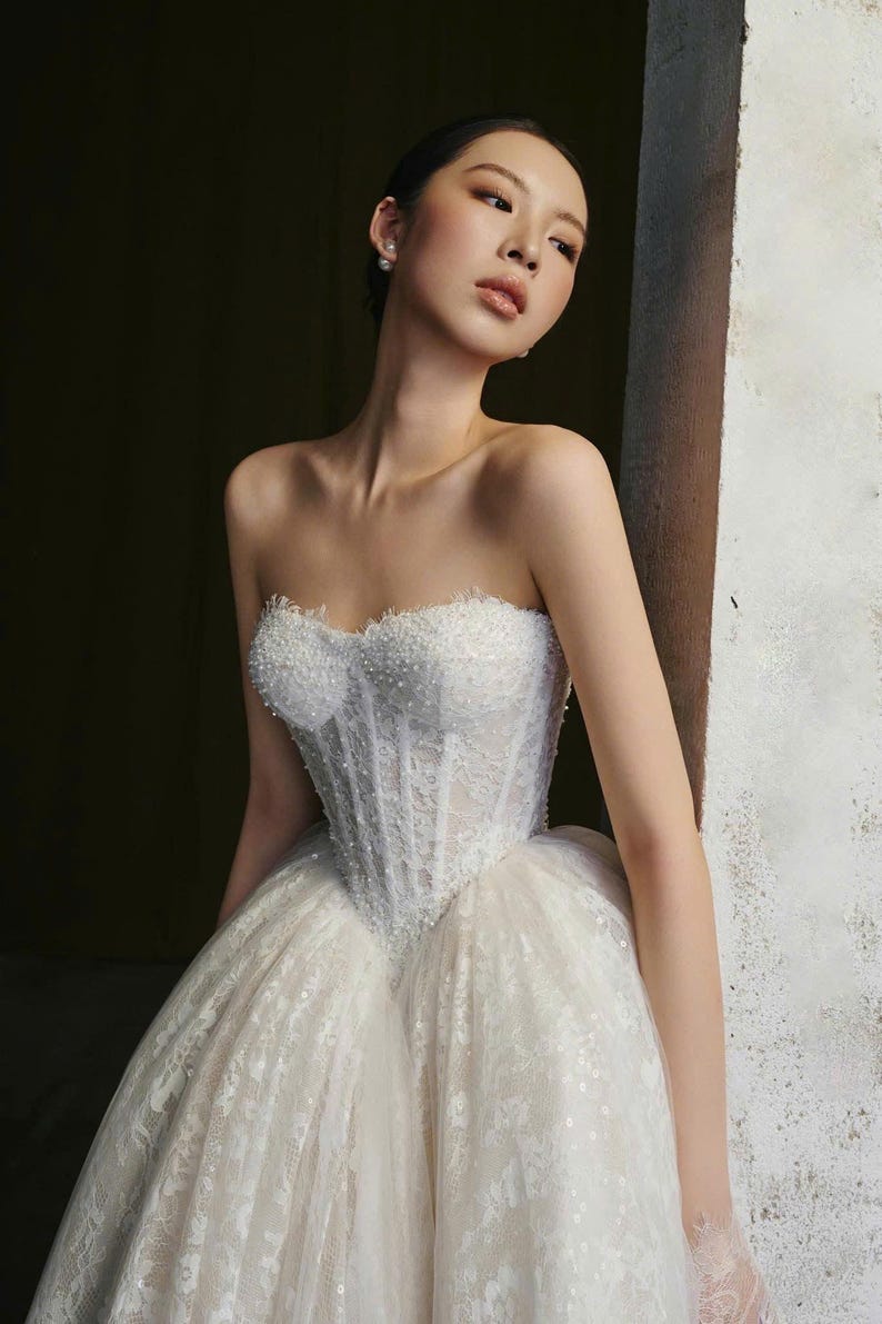 Puede incluir: Un vestido de novia blanco sin tirantes con un corpi&ntilde;o con cuentas y una falda completa y en capas. El vestido presenta intrincados detalles de encaje y un escote coraz&oacute;n. El vestido est&aacute; dise&ntilde;ado para un evento formal.