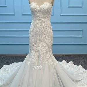 May include: A strapless white wedding dress featuring an intricate lace bodice with a sweetheart neckline and a fitted silhouette that flares into a full tulle skirt with a long train. The number 6491 is printed on the front of the skirt.