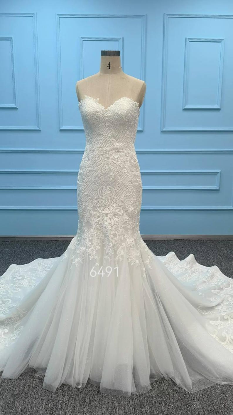 May include: A strapless white wedding dress featuring an intricate lace bodice with a sweetheart neckline and a fitted silhouette that flares into a full tulle skirt with a long train. The number 6491 is printed on the front of the skirt.