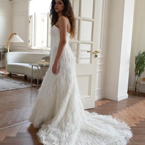 May include: A strapless white floor-length gown featuring a fitted bodice with subtle sequin embellishments and a skirt completely covered in long white feathers that extend into a train, creating a textured and voluminous silhouette.