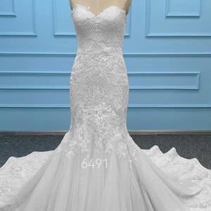 May include: A white strapless mermaid-style gown featuring intricate lace embroidery on the bodice and upper skirt, transitioning into a layered tulle skirt with a long train. The number 6491 is printed in white on the lower front of the dress.