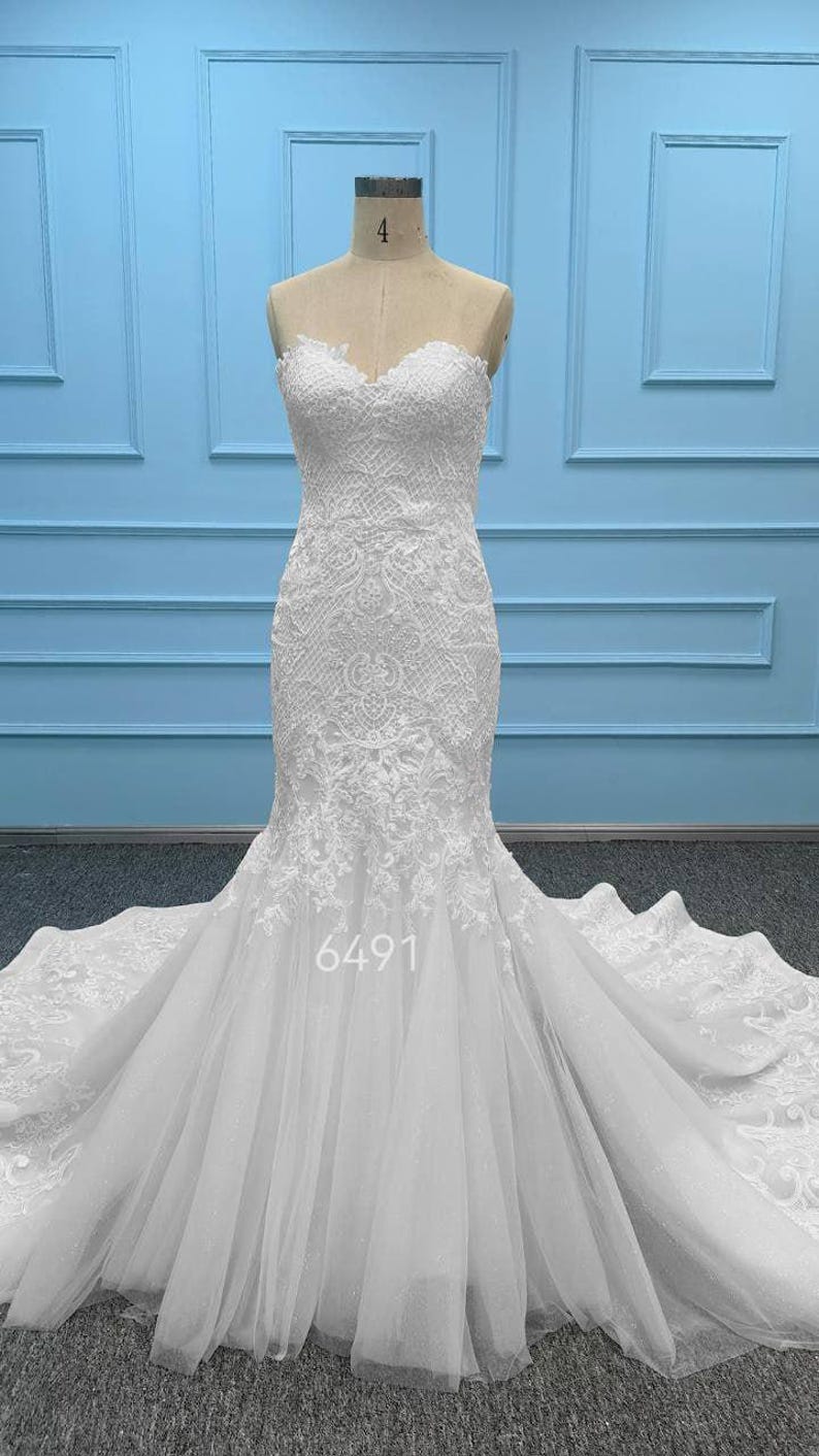 May include: A white strapless mermaid-style gown featuring intricate lace embroidery on the bodice and upper skirt, transitioning into a layered tulle skirt with a long train. The number 6491 is printed in white on the lower front of the dress.