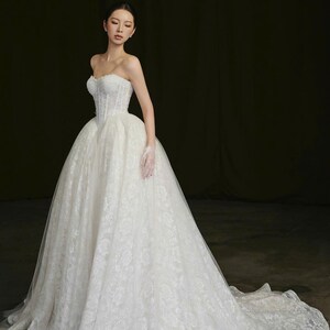 Puede incluir: Un vestido de novia blanco sin tirantes con una falda amplia y una larga cola. El corpi&ntilde;o es ajustado y adornado, y la falda est&aacute; hecha de una delicada tela de encaje. El vestido est&aacute; dise&ntilde;ado para una ocasi&oacute;n formal.