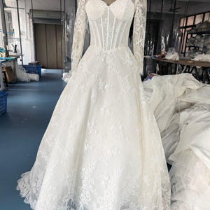 May include: A floor-length white gown featuring long lace sleeves, a sweetheart neckline, and a structured corset-style bodice. The dress is adorned with intricate floral lace overlays that extend from the bodice down to the full, flowing skirt.