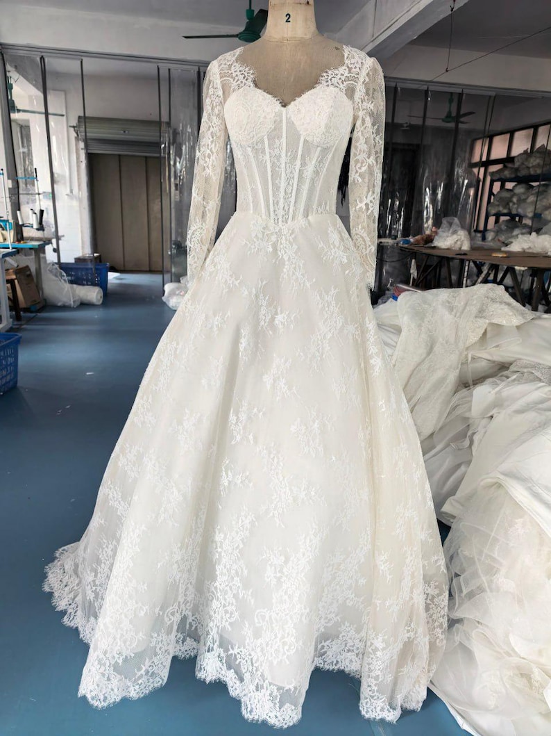 May include: A floor-length white gown featuring long lace sleeves, a sweetheart neckline, and a structured corset-style bodice. The dress is adorned with intricate floral lace overlays that extend from the bodice down to the full, flowing skirt.