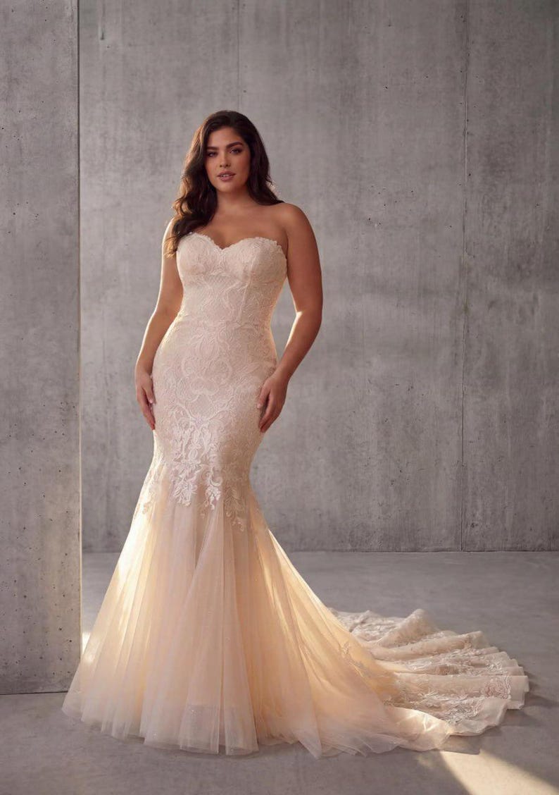 May include: A strapless floor-length gown in a soft champagne hue, featuring a sweetheart neckline and a fitted bodice adorned with intricate white lace embroidery that transitions into a flared mermaid skirt with a long, flowing tulle train.