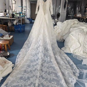 May include: A white floor-length wedding dress with long lace sleeves, a deep V-neckline, and an open back design. The gown features an intricate floral lace overlay that extends into a long, flowing train, showcasing detailed scalloped edges.