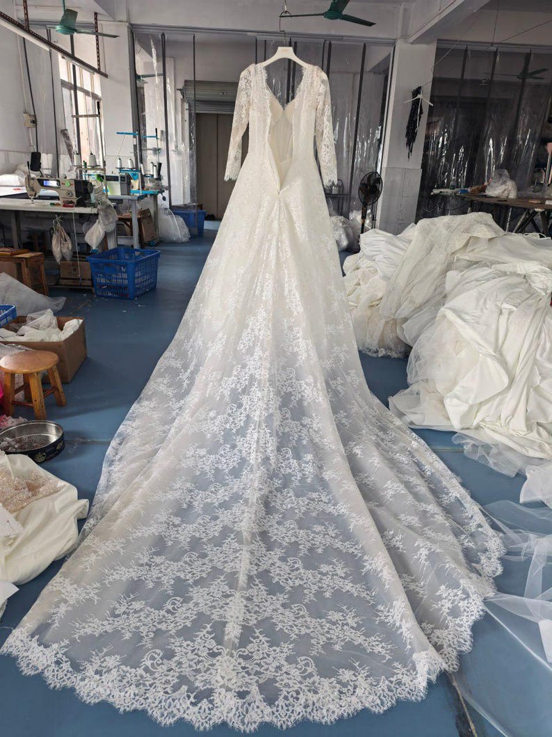 May include: A white floor-length wedding dress with long lace sleeves, a deep V-neckline, and an open back design. The gown features an intricate floral lace overlay that extends into a long, flowing train, showcasing detailed scalloped edges.
