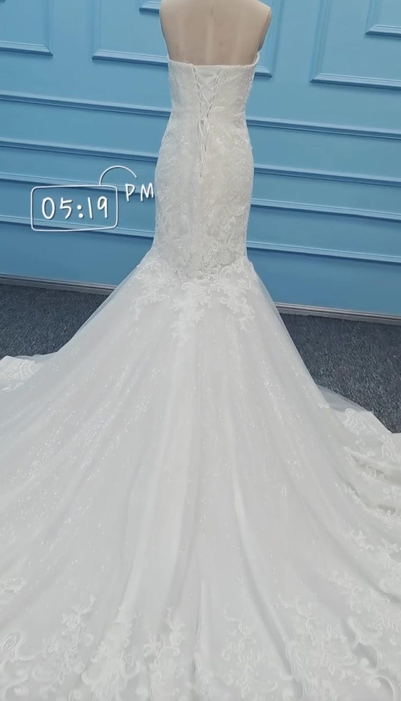 May include: A white strapless floor-length gown featuring a lace bodice with a corset-style lace-up back closure and a flared skirt with a long train, adorned with intricate floral lace appliqu&eacute;s and subtle glitter, displaying the text 05:19 PM.