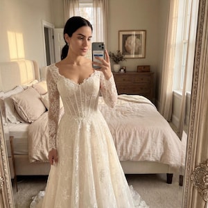 May include: A floor-length ivory wedding dress featuring long lace sleeves, a sweetheart neckline, and a structured corset-style bodice with visible boning. The gown is adorned with intricate floral lace embroidery throughout the skirt and sleeves.
