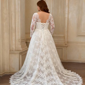 May include: A floor-length white lace gown featuring long sheer sleeves, a deep scoop back, and a corset-style bodice with crisscross lacing. The skirt is full and flows into a train, with intricate floral lace patterns covering the entire garment.