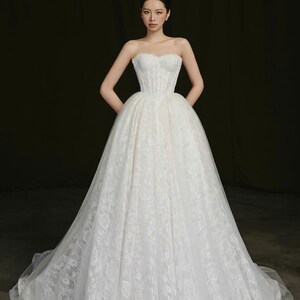 Puede incluir: Un vestido de novia blanco sin tirantes con un corpi&ntilde;o ajustado y una falda amplia y fluida. El vestido est&aacute; adornado con delicado encaje y presenta un escote coraz&oacute;n. La novia est&aacute; de pie con las manos en las caderas.