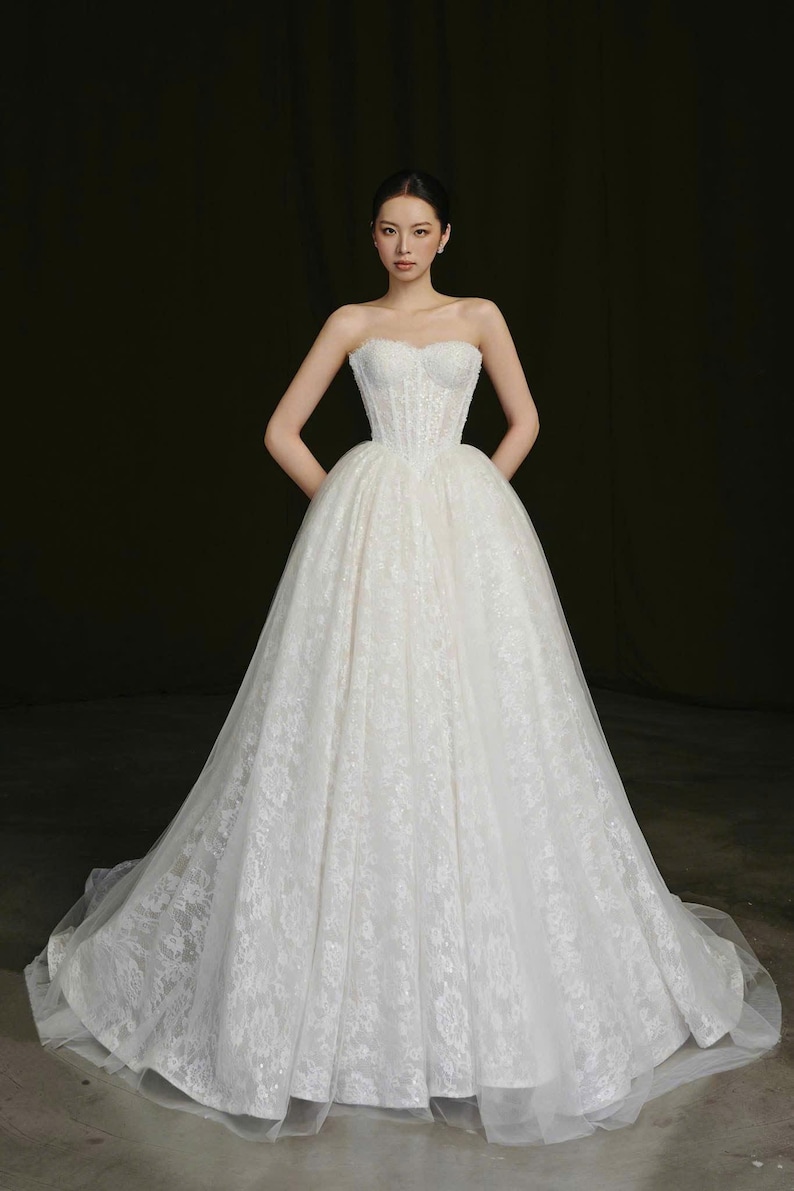 Puede incluir: Un vestido de novia blanco sin tirantes con un corpi&ntilde;o ajustado y una falda amplia y fluida. El vestido est&aacute; adornado con delicado encaje y presenta un escote coraz&oacute;n. La novia est&aacute; de pie con las manos en las caderas.