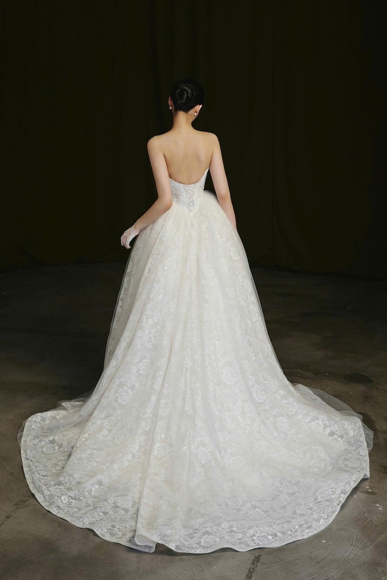 Puede incluir: Un vestido de novia blanco sin tirantes con una falda amplia y una larga cola. El vestido es de encaje y tiene un corpi&ntilde;o ajustado. El pelo de la novia est&aacute; peinado en un mo&ntilde;o.