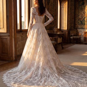 May include: A floor-length white lace gown featuring long sleeves, a corset-style bodice with a lace-up back closure, and a full skirt that extends into a long, intricate lace train with scalloped edges and detailed floral embroidery patterns.