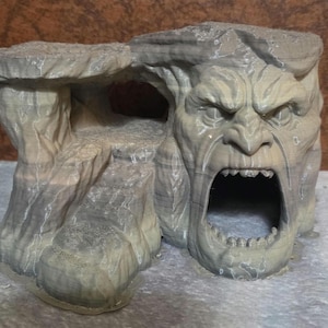 May include: A gray, 3D-printed cave structure with a screaming face carved into the rock. The cave has an open mouth and a small opening on the side. The rock has a textured appearance.