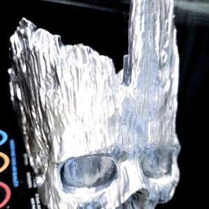 May include: A silver-colored skull sculpture with a textured, wood-like appearance. The skull has large eye sockets and a detailed, rough surface. The sculpture is likely a decorative item or art piece.