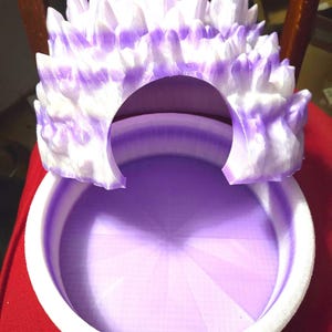 May include: A decorative, 3D-printed object featuring a crown-like structure with a central opening. The crown is white with purple accents, and sits atop a circular, purple-colored base with a white rim. The object is likely a decorative piece or a unique container.