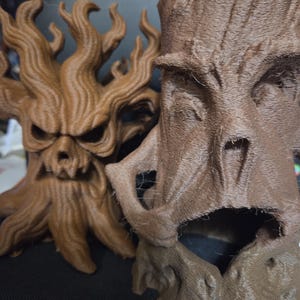 May include: Two brown, 3D-printed tree sculptures with expressive faces. One has a fierce, skull-like appearance, while the other features a more open-mouthed, tree-like face. The sculptures are detailed with textured bark and branches.