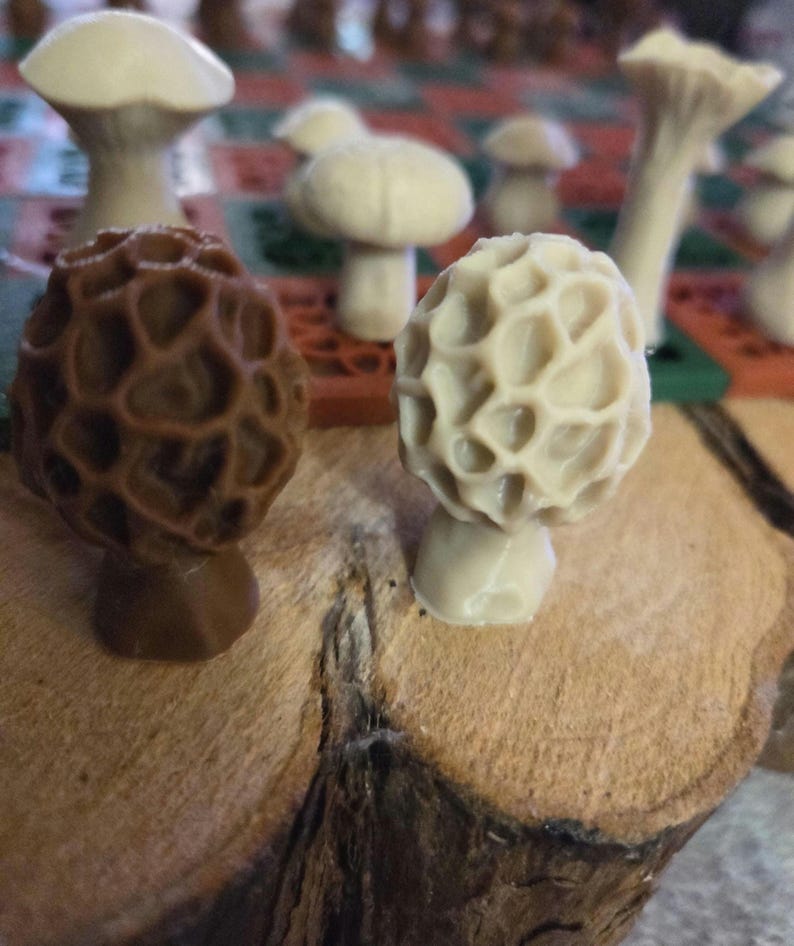 May include: Close-up of several mushroom sculptures, including a brown morel and a cream-colored morel, displayed on a wooden surface. Other mushroom sculptures are visible in the background. The sculptures have detailed textures and varying shapes.