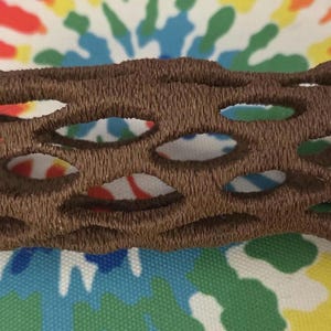 May include: A brown, embroidered cholla cactus skeleton, featuring numerous oval openings. The decorative piece is set against a colorful, tie-dye patterned background, showcasing a natural desert theme.