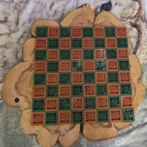 May include: A square, two-tone checkerboard in green and orange, with a floral design in each square. The board is set on a wooden slice with a natural, light brown color and a soft, gray background.