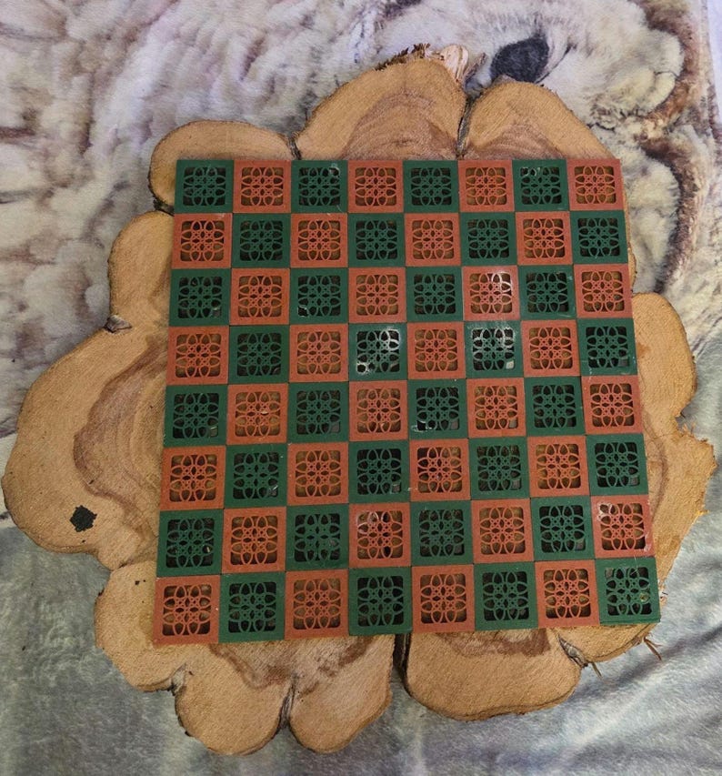 May include: A square, two-tone checkerboard in green and orange, with a floral design in each square. The board is set on a wooden slice with a natural, light brown color and a soft, gray background.