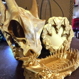 May include: Gold-colored dragon skull and jawbone, with a matching circular base. The skull has detailed texture and visible eye sockets. The jawbone features sharp teeth. The items appear to be 3D printed.
